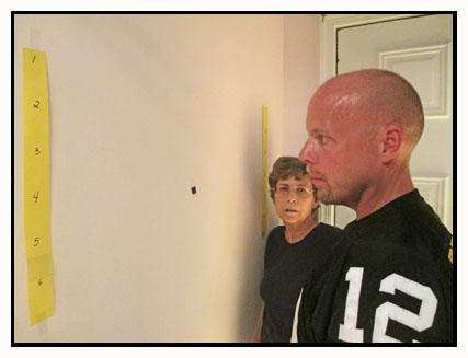 Two photos show the wall with a black spot between two yellow numbered strips.  The man in the football jersey is looking to the left and then looks forward and points to the black spot, as Dona watches.